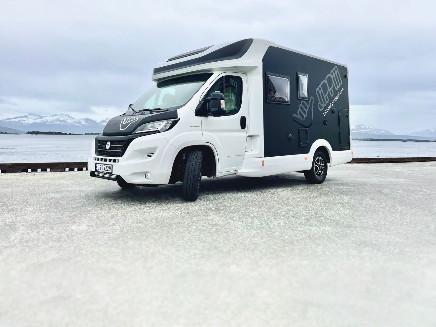 Jippiii-bobilen parkert ved fjorden med snødekte fjell i bakgrunnen